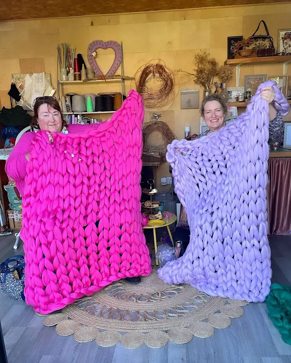 Two women holding up blankets knitted at an event.